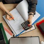 A professional reviewing general ledger transactions on a dual-screen monitor with financial charts.